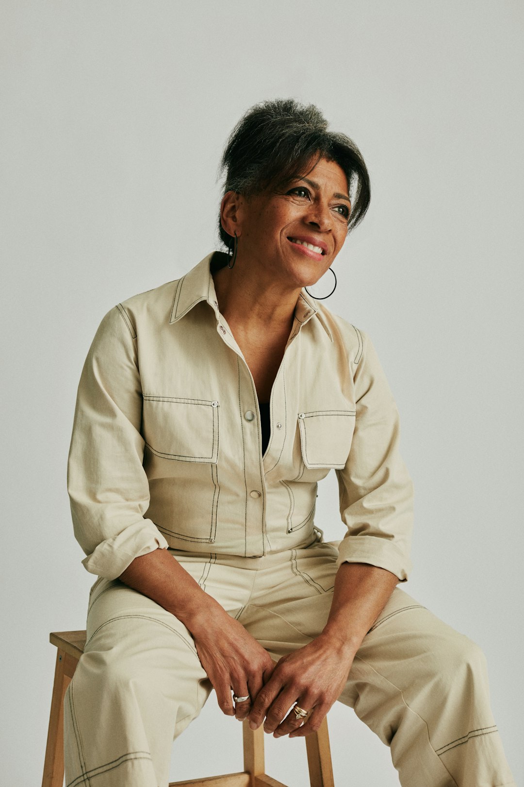 Woman in beige jumpsuit smiling, sitting on a stool